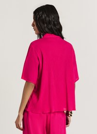 Chemise oversize fuchsia à manches courtes, texture plissée et col. Le dos est légèrement plus long, ajoutant fluidité et dimension.