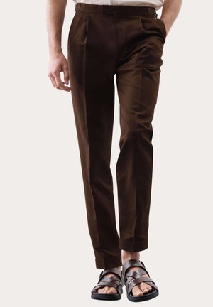 Hombre con pantalones ajustados marrón oscuro y sandalias de cuero marrón oscuro, de pie frente a un fondo claro y liso.