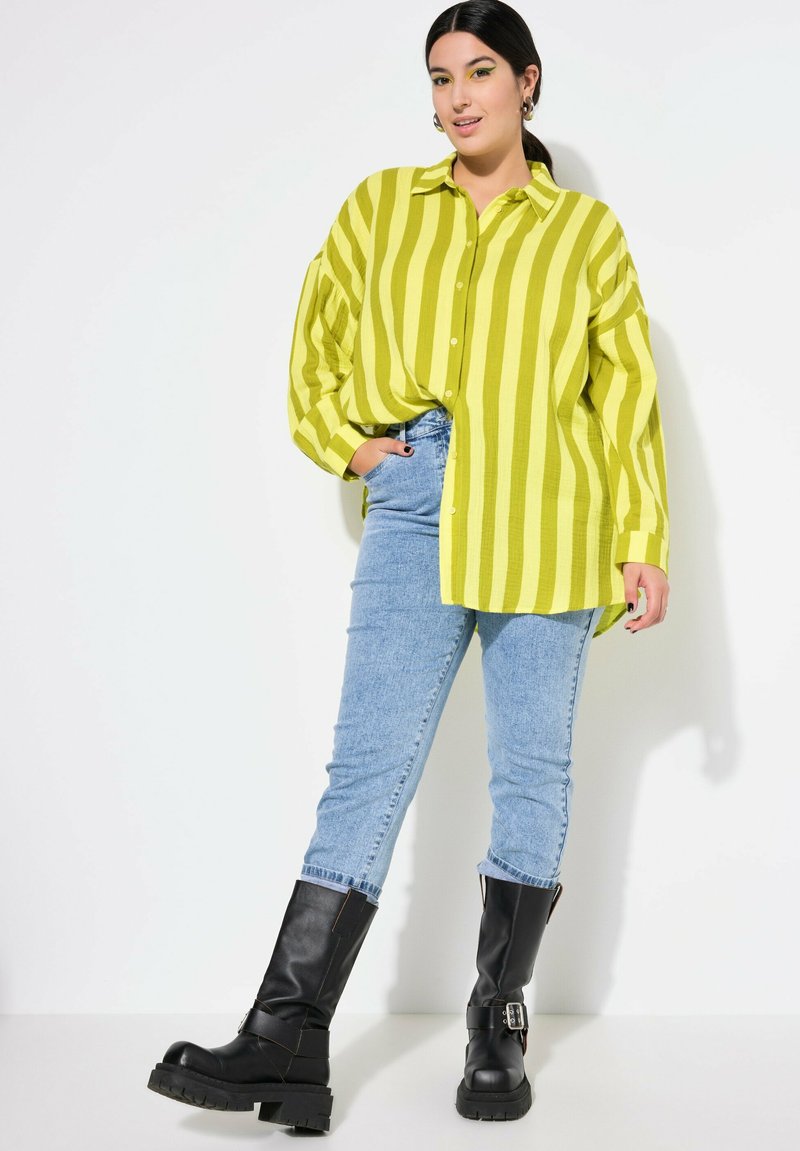 Camisa de manga larga con botones en un vibrante verde lima, con audaces rayas amarillas; combinada con jeans ajustados azul claro y botas negras hasta la rodilla.