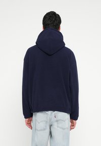 Temno modra jopa z veliko nastavljivo kapuco, sproščena kroja in rebrastim robom. Kombinirana s svetlo modrimi hlačami iz jeansa z logotipom na zadnjem žepu.