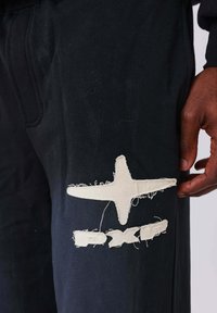 Pantalones de chándal negros de algodón con costuras visibles y parches de tela beige en forma de estrella y texto en la zona del muslo.