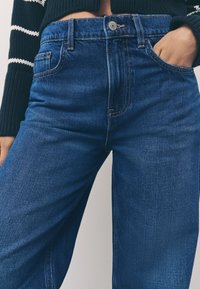 Tumši zilas džinsa bikses ar taisnu piegriezumu, kurām ir rāvējslēdzēja priekšpoga, priekšējās kabatas un redzamas šūšanas detaļas. Auduma viegli izteikta tekstūra.