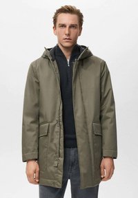 Parka impermeable verde oliva con capucha, cierre frontal de cremallera, dos bolsillos de parche y corte recto. Lleva encima un suéter negro texturizado.