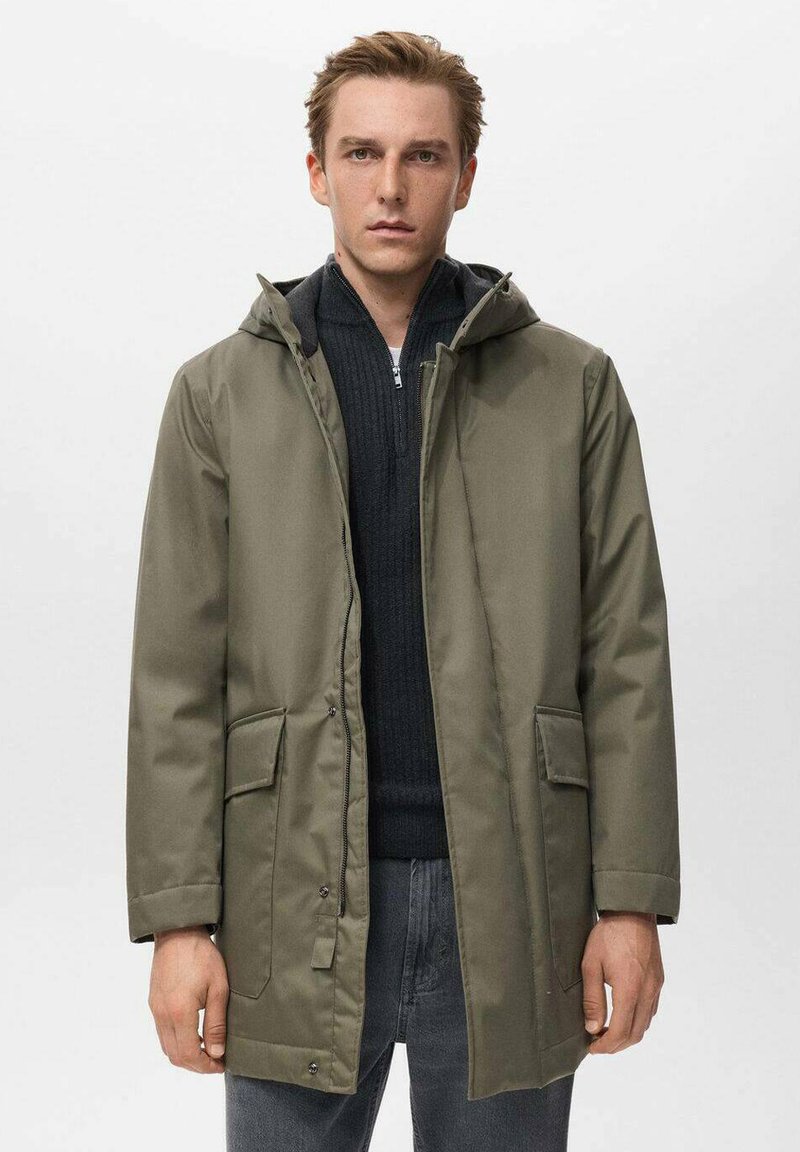 Parka impermeable verde oliva con capucha, cierre frontal de cremallera, dos bolsillos de parche y corte recto. Lleva encima un suéter negro texturizado.