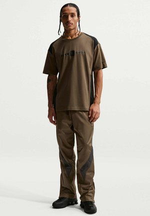 Uomo con capelli intrecciati che indossa una maglietta Nike Shox marrone e nera, pantaloni coordinati e sneakers nere, in piedi contro uno sfondo neutro.