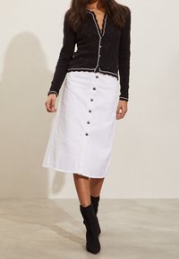 Haut noir à manches longues avec des bords festonnés, associé à une jupe blanche à boutons à l'avant et des bottines noires. Tissu lisse et design ajusté.