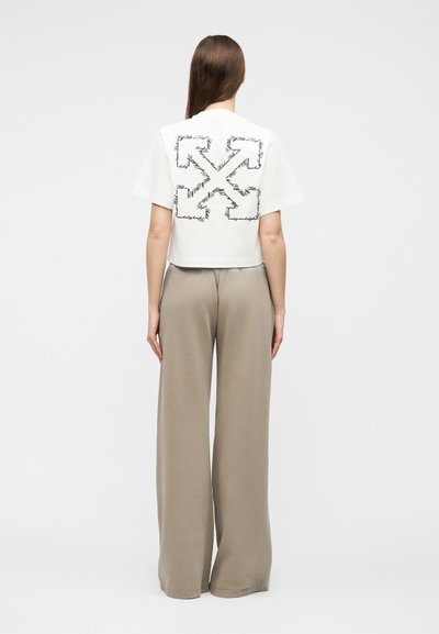 Γυναίκα με μακριά μαλλιά που φοράει λευκό κοντό μπλουζάκι με μαύρο σχέδιο βέλους και μπεζ παντελόνι φαρδύ ποδόγυρο, στέκεται με την πλάτη προς την κάμερα.