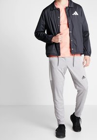 Chaqueta ligera negra con botones a presión y un logo blanco, combinada con una camiseta rosa y pantalones joggers grises con acentos laterales. Zapatillas negras.