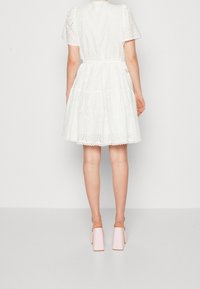 Robe en coton blanc à manches courtes, motif broderie anglaise, jupe froncée et ceinture nouée à la taille. Portée avec des mules à talons épais rose pâle.