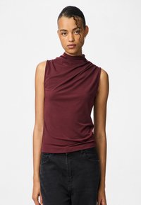 Top burdeos sin mangas con escote drapeado y suavidad en la textura, combinado con jeans negros. Presenta un diseño ajustado sin herrajes visibles.