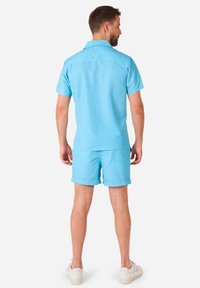 Camicetta azzurra a maniche corte con colletto abbinata a pantaloni corti dello stesso colore, realizzata in tessuto morbido, con bordi arrotolati, indossata con sneakers bianche.