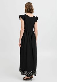 Robe maxi noire avec des manches à volants, taille froncée et broderie de feuilles blanches à l'ourlet. Le dos présente un col rond et une fermeture éclair.