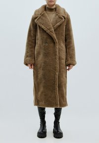 Brauner Kunstfellmantel mit Schalkragen, doppelt geknöpftem Design und Seitentaschen, kombiniert mit knielangen schwarzen Plateau-Stiefeln.