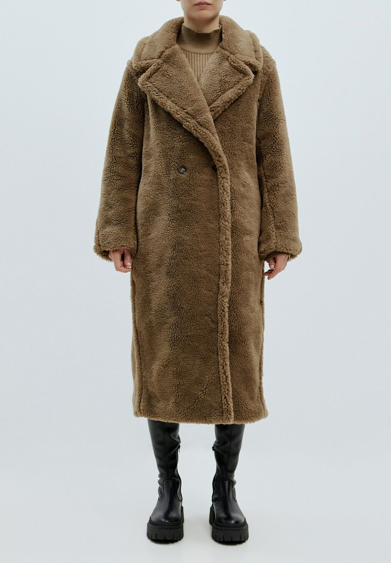 Brauner Kunstfellmantel mit Schalkragen, doppelt geknöpftem Design und Seitentaschen, kombiniert mit knielangen schwarzen Plateau-Stiefeln.