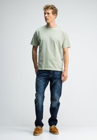 Lichtgroene katoenen t-shirt met korte mouwen en een klein logo op de borst, gecombineerd met donkerblauwe denim jeans en bruine suède schoenen.