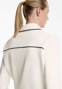 White blazer featuring black piping along the back, tailored fit, and smooth texture, with a notched collar and minimalistic design.