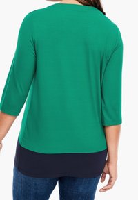 Maglietta verde con maniche a tre quarti, caratterizzata da un pannello inferiore blu navy. Tessuto morbido con una trama liscia. L'orlo è leggermente curvato.