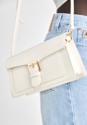 White leather crossbody bag with gold buckle worn over light blue denim jeans against a plain white background.