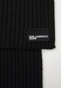 Cachecol de malha canelada preto com um rótulo cosido a dizer "KARL LAGERFELD JEANS". Apresenta uma trama texturizada e uma forma retangular.