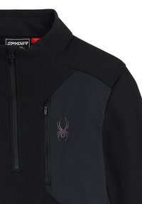 Schwarze Fleecejacke mit strukturierten Strick-Schultern, Reißverschlusstasche und lila Spinnenlogo auf der Brust. Glattes Gewebe mit kontrastierendem Design.