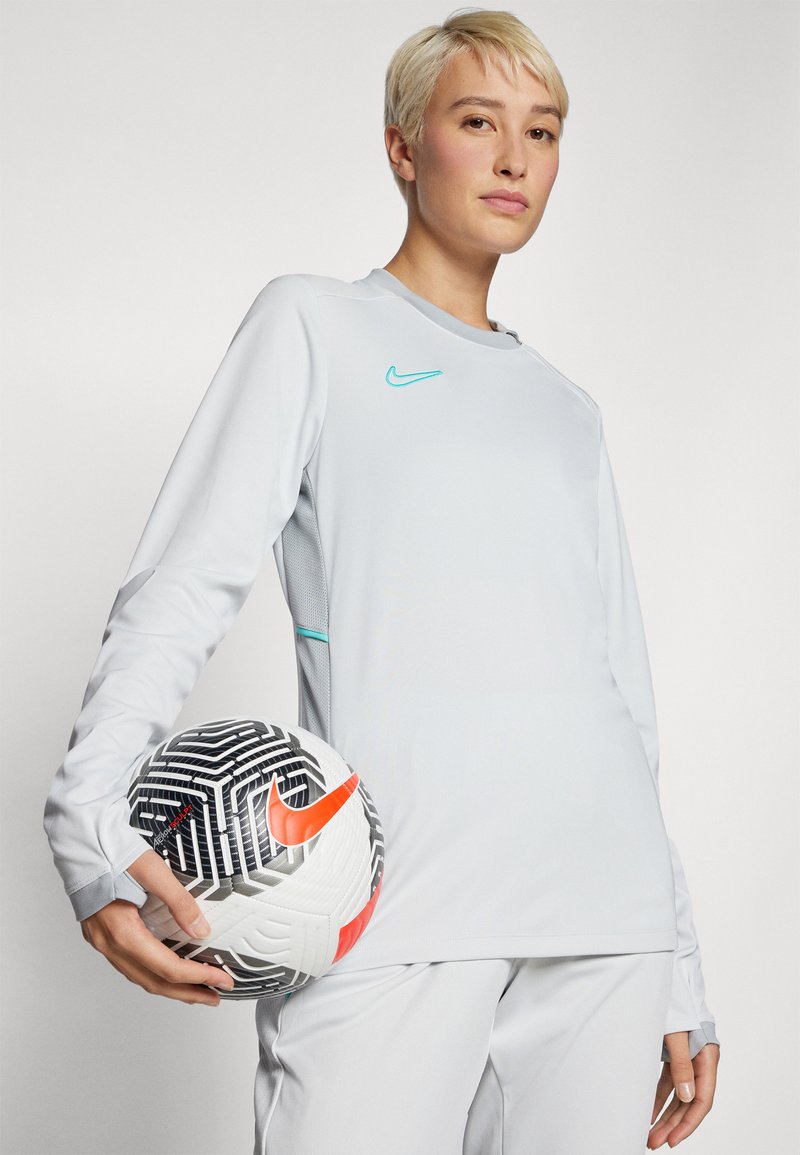 Lange mouw, lichtgrijze sporttop met turkoois accent. Speler houdt een gemêleerde zwart-witte voetbal met oranje logo vast.