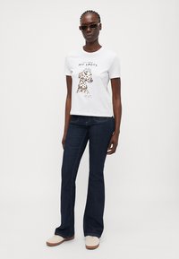 White cotton t-shirt featuring a printed dog graphic and text, paired with dark blue flared jeans and beige footwear.