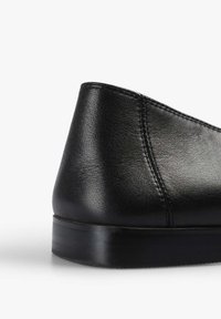Zapato de cuero negro con punta redondeada y un tacón bajo y liso. La superficie presenta una textura sutil, con costuras visibles a lo largo de las uniones.