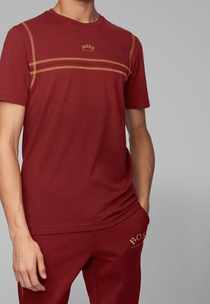 Homme portant un t-shirt rouge à manches courtes avec des coutures jaunes et un pantalon assorti, tous deux affichant le logo "BOSS Hugo Boss".