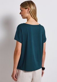 Teal kort mouwen top met een wijde halslijn en een relaxte pasvorm; gladde textuur en subtiele glans. Gestyled met lichte linnen broek.