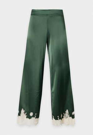 Kobieta w ciemnozielonej satynowej koszulce na ramiączkach i szerokich spodniach z białymi koronkowymi detalami, połączona z czarnymi sandałami na obcasie, stojąca na jednolitym tle.