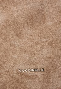Taupe zamška z gladko teksturo; blagovna znamka "COCCINELLE" je upodobljena v kovinsko embosirani pisavi na dnu.