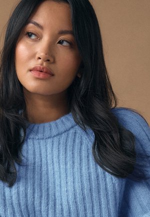 Jauna sieviete ar gariem melniem matiem, ģērbusies gaiši zilā ribota džempera, domīgi skatās uz sāniem uz neitrāla fona.