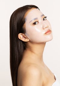 Gennemsigtig ansigtmaske anvendt på ansigtet, glat struktur, gennemskinneligt materiale, med udskæringer til øjne og mund, mod en neutral baggrund.