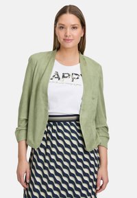 Blazer vert clair en suède, de coupe décontractée et avec des manches retroussées, porté sur une chemise blanche et une jupe à motifs bleu marine et crème.