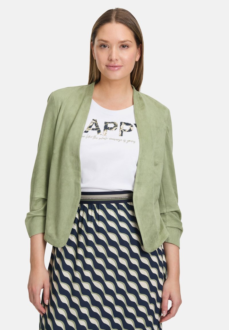 Blazer vert clair en suède, de coupe décontractée et avec des manches retroussées, porté sur une chemise blanche et une jupe à motifs bleu marine et crème.