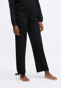 Črne trenirke s sproščenim krojem, z elastičnimi manšetami in gladko teksturo tkanine. Oblikovane z vrvico na spodnjem robu.