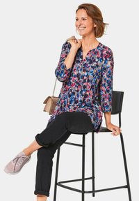 Bunte Blumenbluse mit Dreiviertelärmeln, getragen über hochgekrempelten schwarzen Hosen und rosa Sneakers. Die Bluse hat einen V-Ausschnitt und Falten.