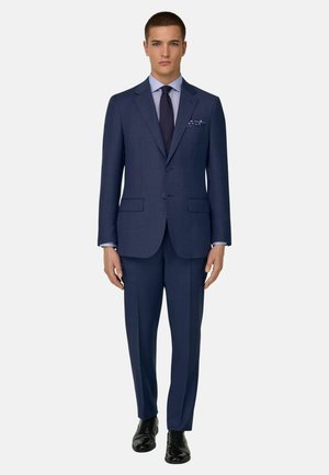 Jonge man staand gekleed in een op maat gemaakt marineblauw pak met bijpassende broek, lichtblauw overhemd, donkere stropdas, met patroon versierde pochet en zwarte nette schoenen.