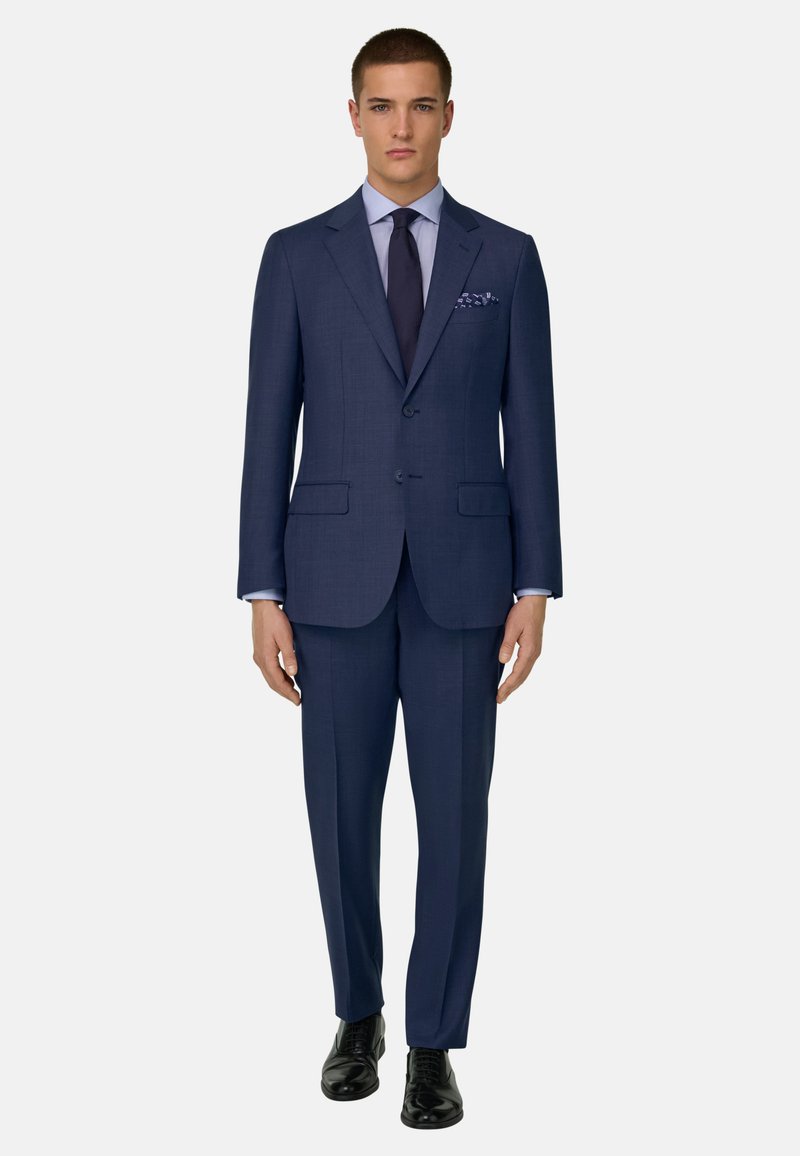 Jeune homme debout portant un costume bleu marine sur mesure avec un pantalon assorti, une chemise bleu clair, une cravate sombre, un mouchoir de poche à motif et des chaussures habillées noires.