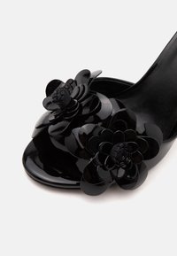 Sandale à talon haut ouverte à l’avant en cuir verni noir avec deux applications florales décoratives superposées sur la bride avant.