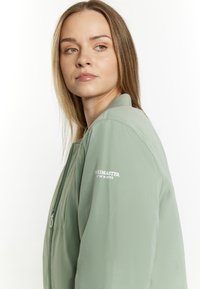 Lichtgroene bomberjack met een ritssluiting, geribbelde manchetten en een klein geborduurd logo op de borst. Zachte, gladde stoftextuur.