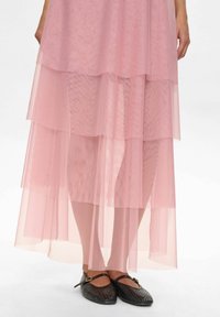 Piernas inferiores con una falda de tul transparente en capas de color rosa, combinadas con unas bailarinas negras de estilo Mary Jane con correas de hebilla sobre un fondo blanco.