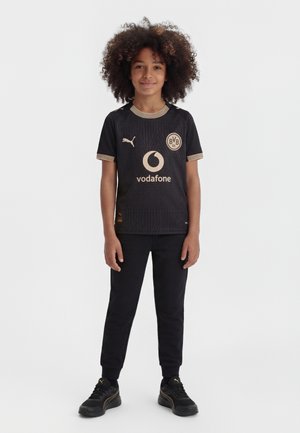 Dieťa s kučeravými vlasmi, oblečené v čiernom futbalovom drese Borussia Dortmund, čiernych nohaviciach a čiernych teniskách Puma, stojace pred bielym pozadím.