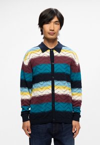 Cardigan tricoté multicolore avec des motifs en zigzag dans des nuances de bleu, rouge, jaune et blanc, avec un col sombre et une fermeture à boutons.
