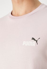 Camiseta de manga corta color rosa claro, hecha de una tela suave, con un logo negro de Puma impreso en el área del pecho izquierdo. Cuello redondo.