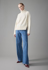 Pull à col roulé en crème avec ourlet côtelé, associé à un jean en denim bleu ample. Pieds dans des escarpins bordeaux à bout pointu, texture lisse et coupe décontractée.