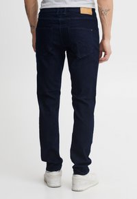 Sötétkék farmernadrág slim fit szabással és két hátsó zsebbel. A derekán bézs bőr folt található. Fehér sportcipővel viselve.