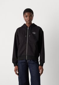 Black zip-up hoodie with front pockets, featuring a small Calvin Klein logo. The fabric appears soft with a smooth texture and relaxed fit.