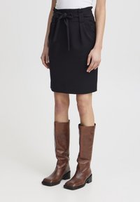 Jupe noire mi-longue avec une ceinture nouée, en tissu lisse. Associée à des bottes en cuir marron montant jusqu'aux genoux avec un talon épais.