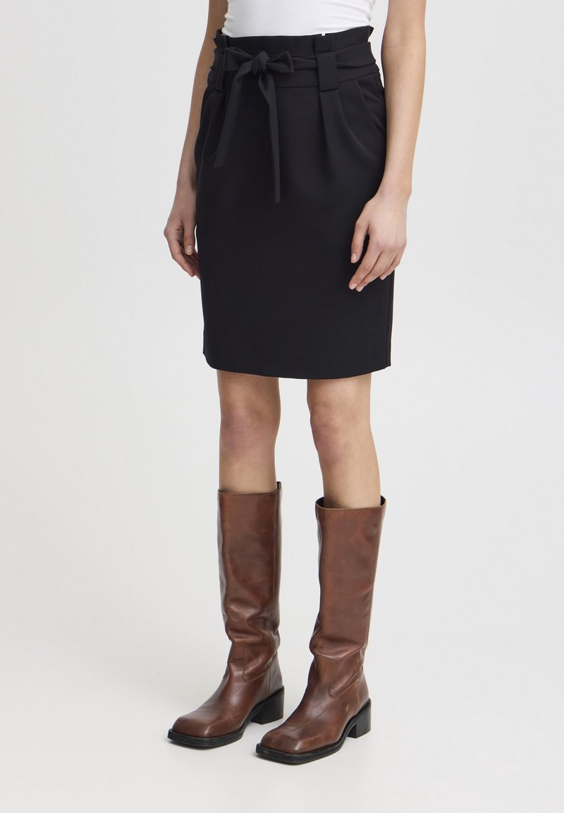 Jupe noire mi-longue avec une ceinture nouée, en tissu lisse. Associée à des bottes en cuir marron montant jusqu'aux genoux avec un talon épais.